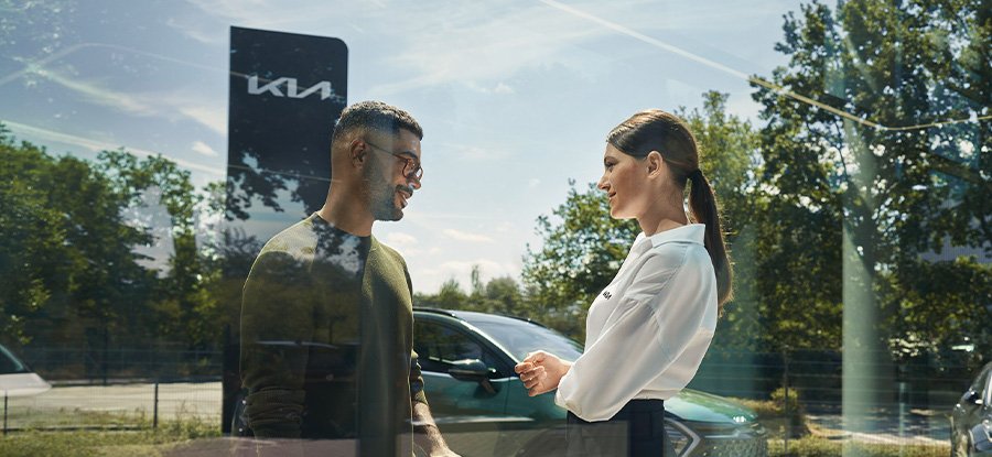 Ein Mann und eine Frau sprechen lächelnd vor einem Kia-Auto in einer sonnigen Umgebung.