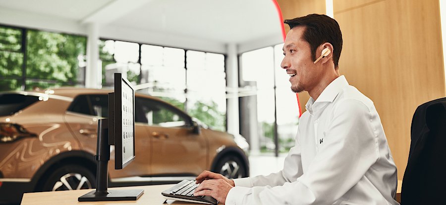 Ein Berater in einem Autohaus sitzt lächelnd am Computer mit Kopfhörern. Im Hintergrund ist ein Auto zu sehen.