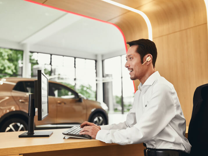 Ein Mann mit Kopfhörern arbeitet an einem Computer in einem modernen Autohaus mit einem Auto im Hintergrund.