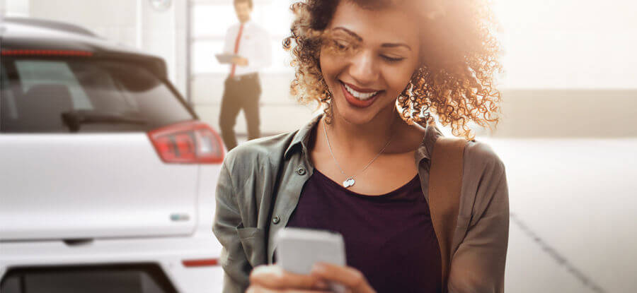 Eine lächelnde Frau mit Locken schaut auf ihr Handy, während ein Mann im Hintergrund steht und einen Anzug trägt.