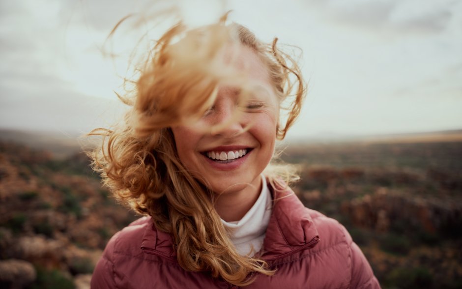Lächelnde Frau mit wehendem Haar, die vor einer Landschaft steht, im Freien, fröhliche Atmosphäre.