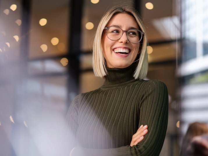 Lächelnde Frau mit Brille und blonden Haaren, trägt einen grünen Rollkragenpullover in einem modernen Raum.