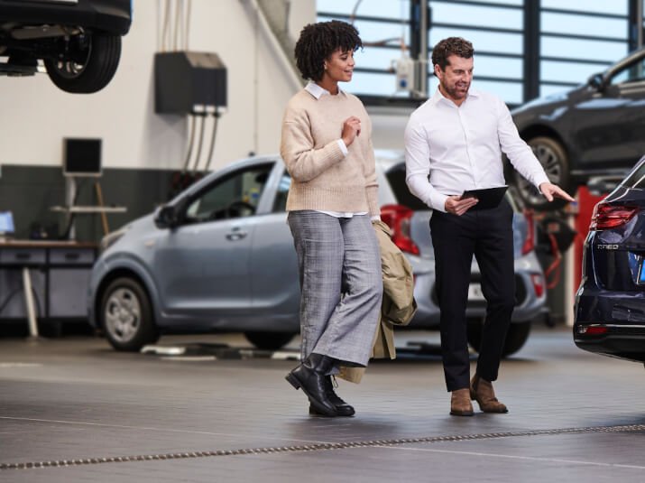 Eine Frau und ein Mann besprechen ein Auto in einer Werkstatt, umgeben von weiteren Fahrzeugen.