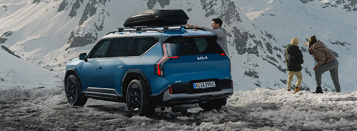 Ein blauer Kia SUV in den Bergen, mit Schnee und drei Personen, die die Aussicht genießen.