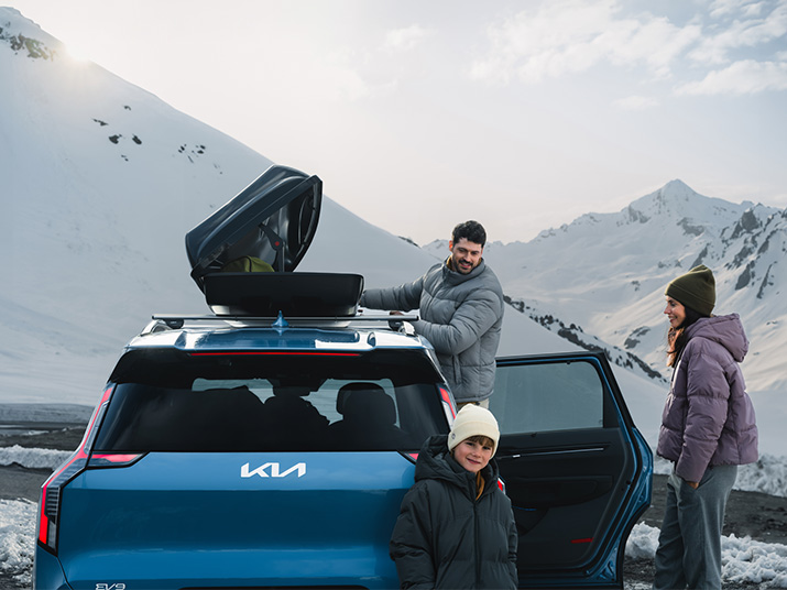 Eine Familie steht an einem Kofferraum eines Kia Fahrzeugs in den Bergen, umgeben von Schnee.